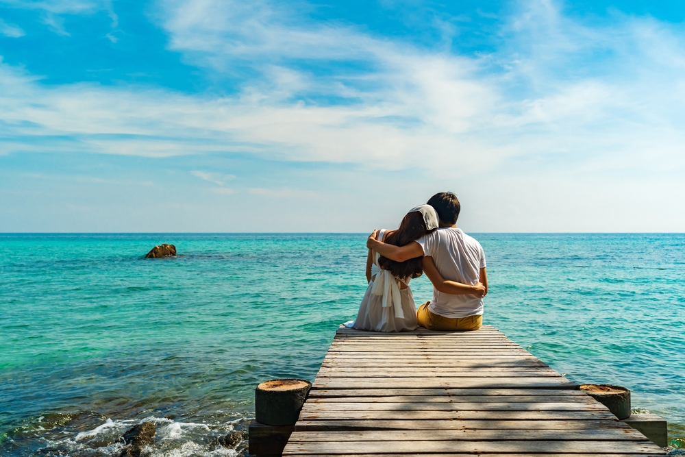 A couple embracing on a wooden pier during their Thailand honeymoon trip, looking out at the calm turquoise ocean under a blue sky