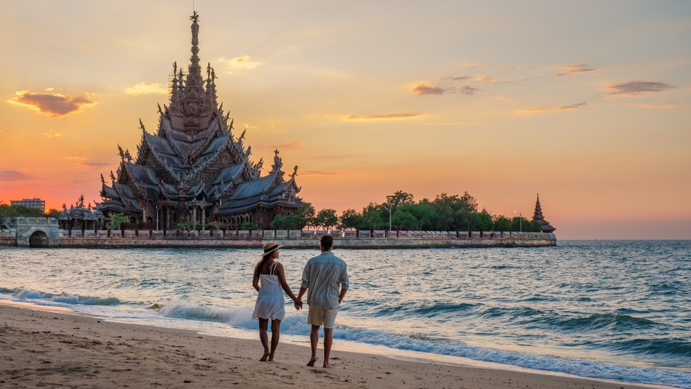 Couple walking on a beach at sunset with the Sanctuary of Truth in the background, perfect for a romantic Thailand honeymoon.