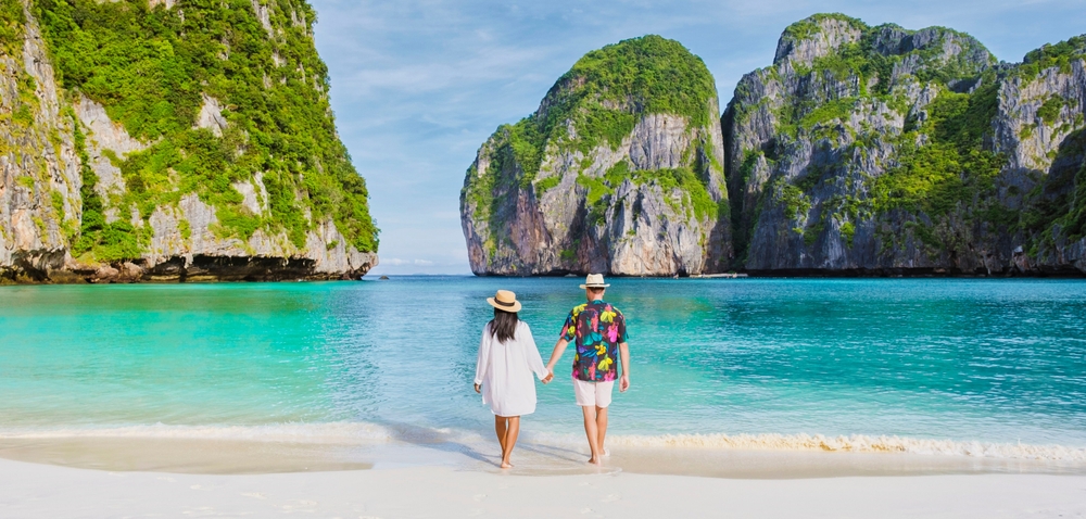 A couple on their Thailand honeymoon trip walking hand-in-hand on a white sand beach with turquoise water and limestone karsts at Maya Bay, Phi Phi Islands 