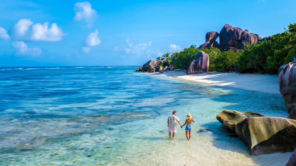 A couple walking hand-in-hand in shallow turquoise water during a Seychelles or Maldives honeymoon at Anse Source d'Argent beach with iconic granite boulders