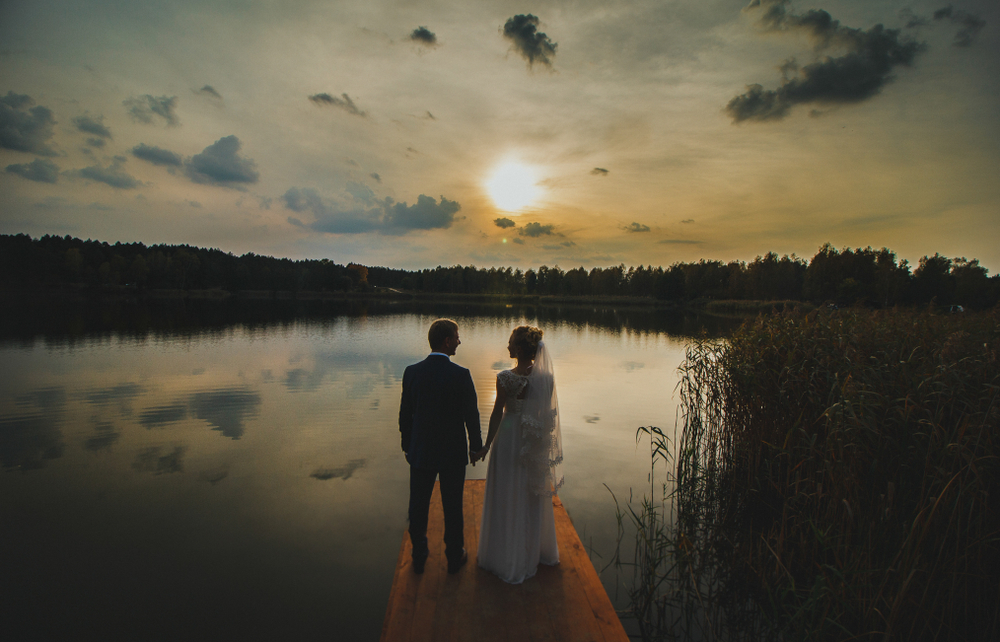 A newly married couple enjoying a Ninh Bình honeymoon, standing hand-in-hand on a wooden pier at sunset overlooking a tranquil lake surrounded by lush forests.