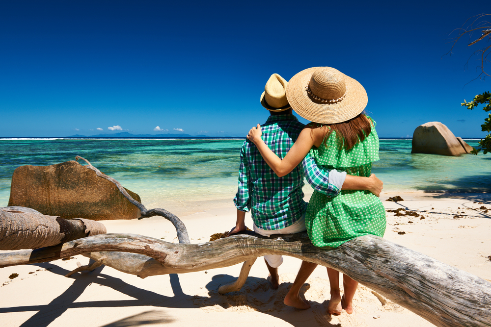 Rear view of a man and woman in green summer attire sitting on a driftwood log, overlooking the Indian Ocean and distant islands during a Seychelles honeymoon trip. 