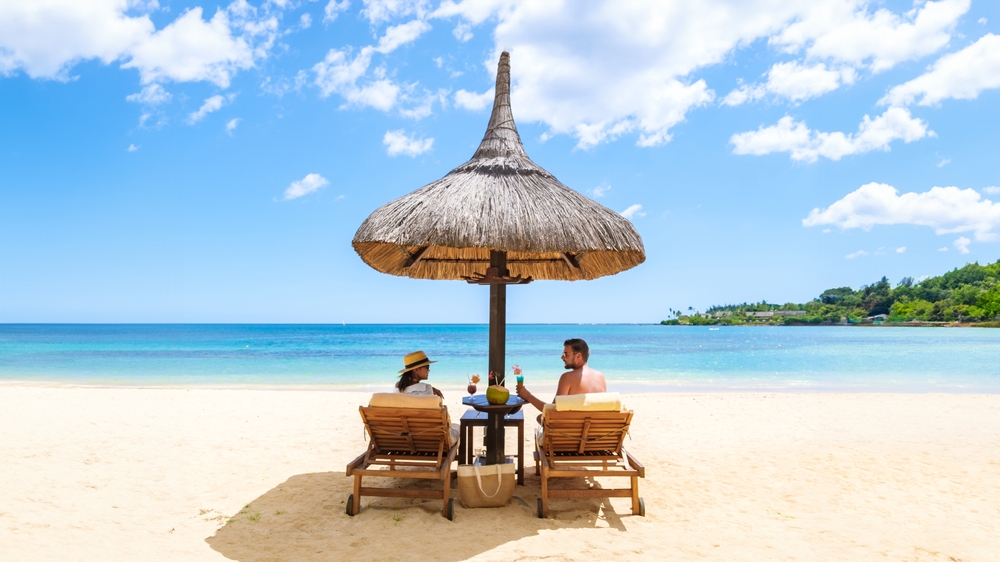 A couple enjoying a Mauritius honeymoon under a thatched beach umbrella on a white sand beach with turquoise ocean water.