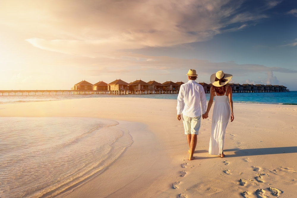 A couple holding hands in shallow turquoise water on a white sand beach during their Maldives island honeymoon, surrounded by unique granite boulders and lush tropical greenery