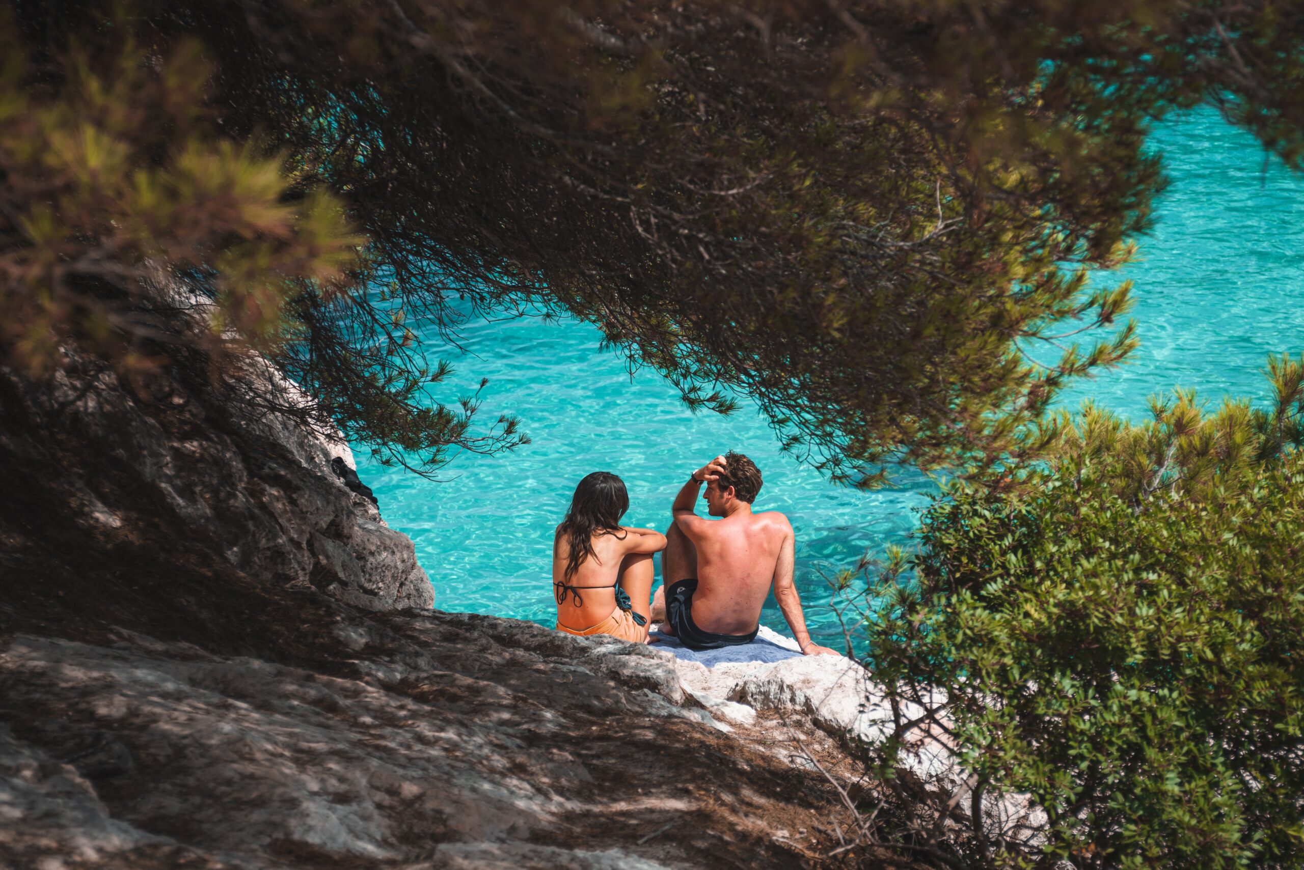 A romantic couple sitting on a shaded rocky shore overlooking the crystal-clear turquoise ocean during their Seychelles honeymoon.