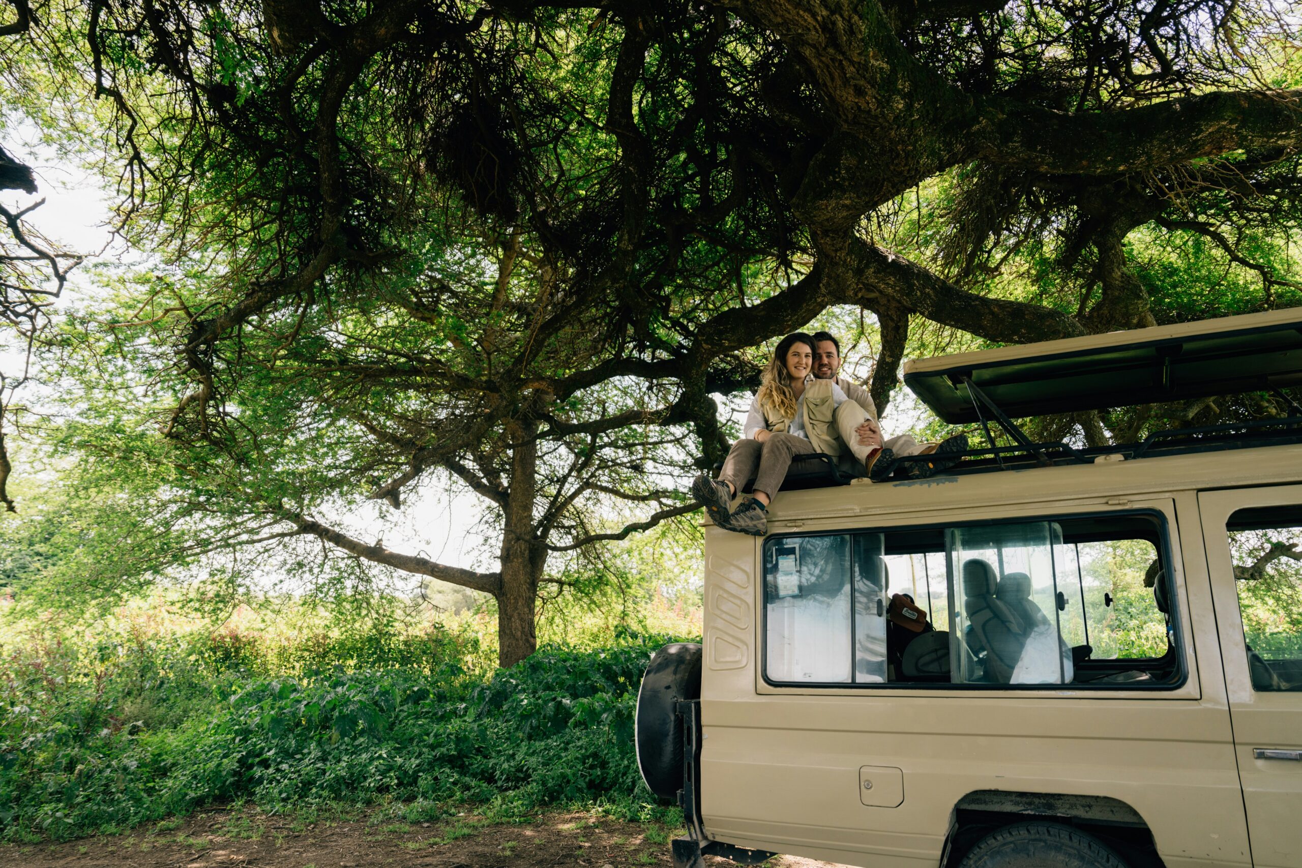 A smiling couple enjoys the view of the savanna landscape from the roof of their safari Land Cruiser parked under a massive acacia tree during a tanzania honeymoon safari.