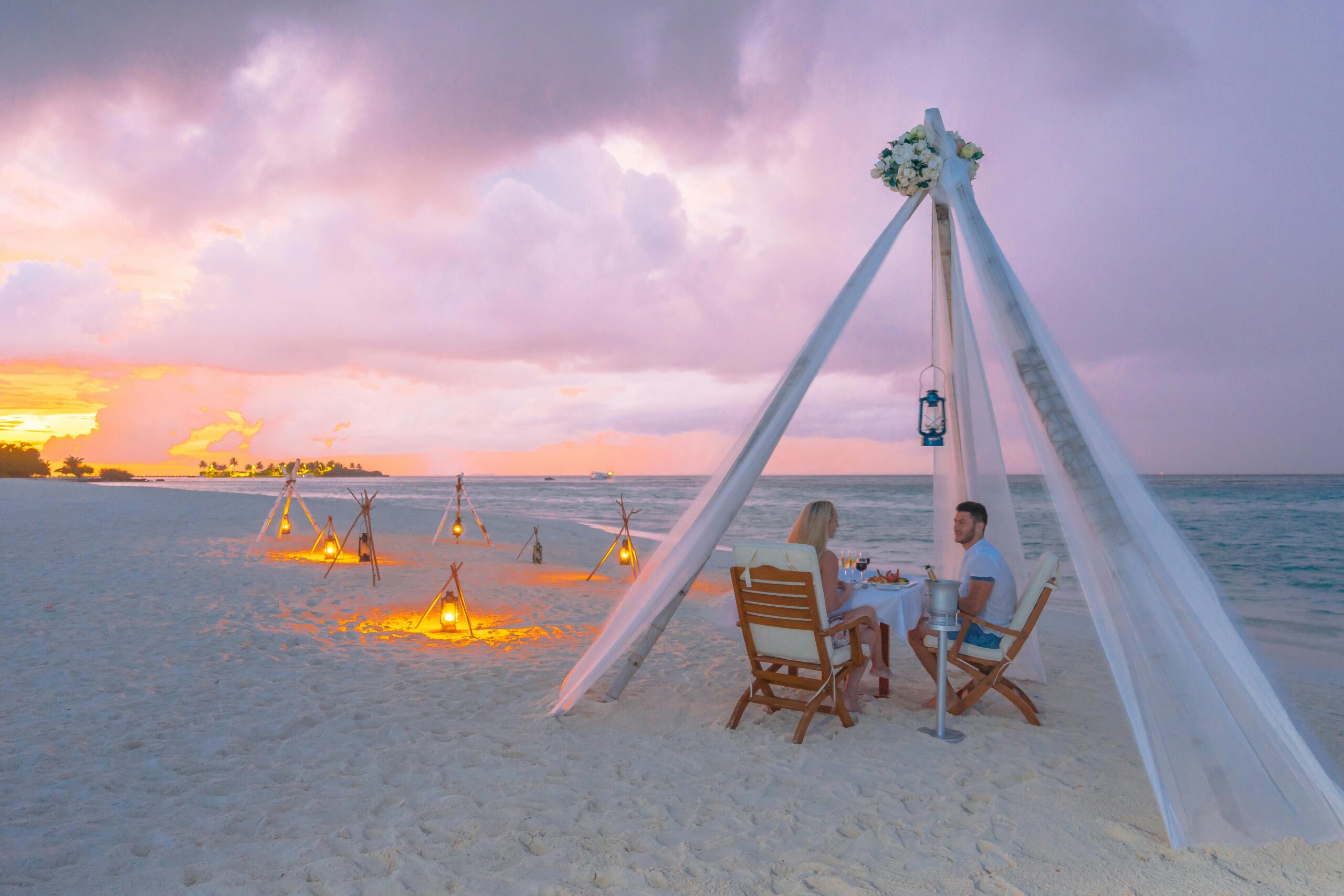 A couple enjoying a private beach glamping dinner at sunset under a white canopy with glowing lanterns, showcasing one of the most romantic date ideas singapore has to offer.
