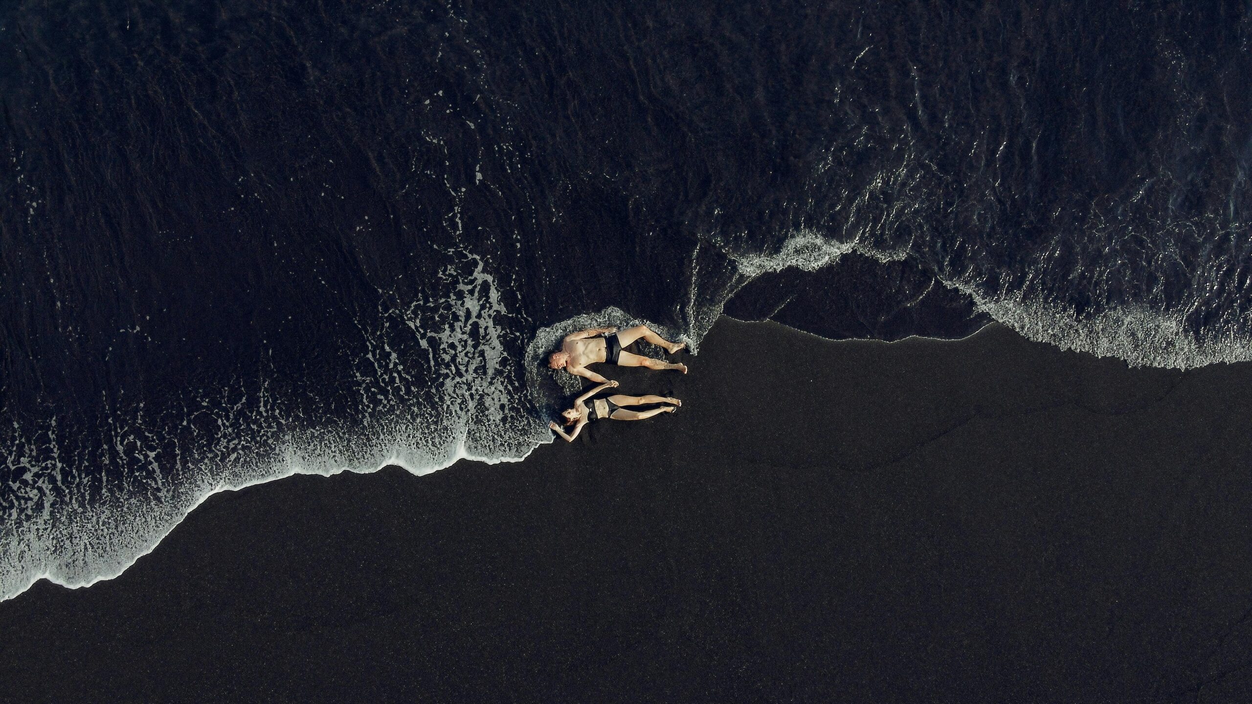 Best area to stay in bali for couples: An aerial view of a romantic couple holding hands and relaxing on a dramatic black sand beach with ocean waves.