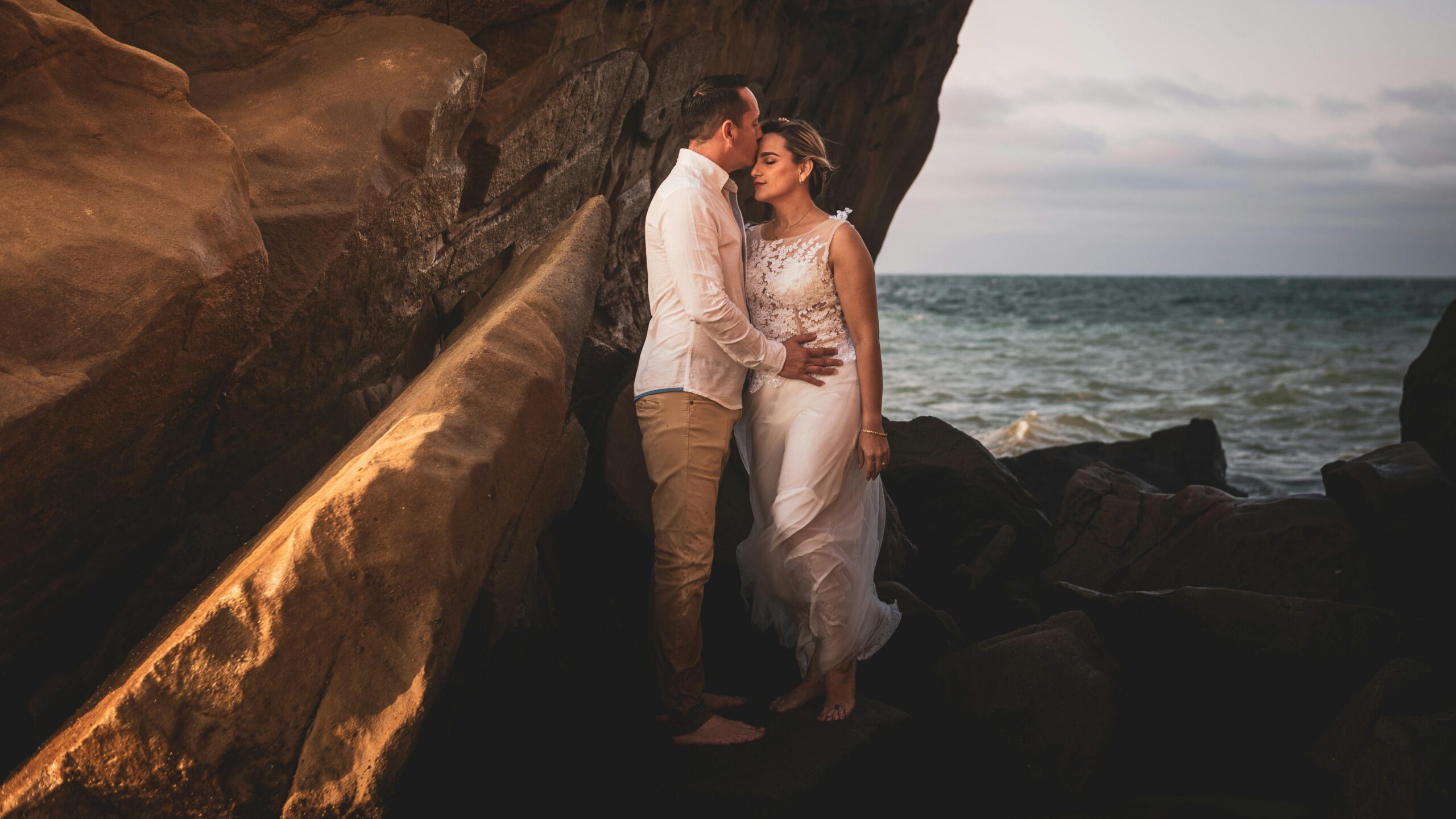 An intimate portrait of a couple, with the bride in a white lace wedding dress and the groom kissing her forehead, standing among coastal rocks by the sea, capturing a romantic moment on their Seychelles Honeymoon.