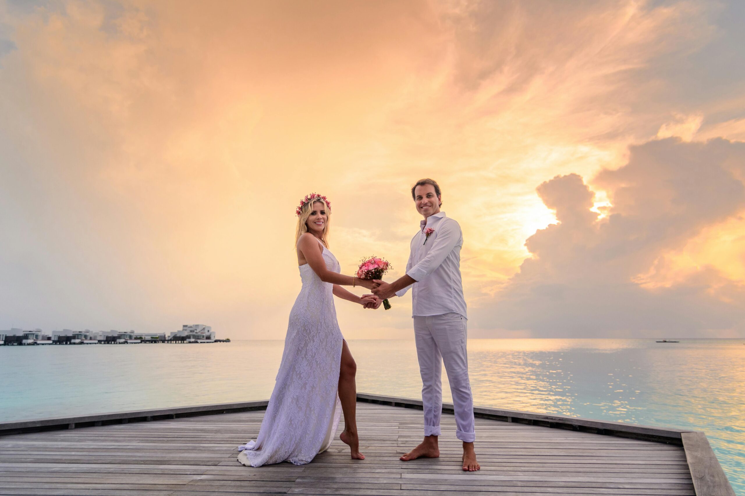 A romantic newlywed couple on a wooden pier at sunset with overwater villas, experiencing the best maldives honeymoon travel packages