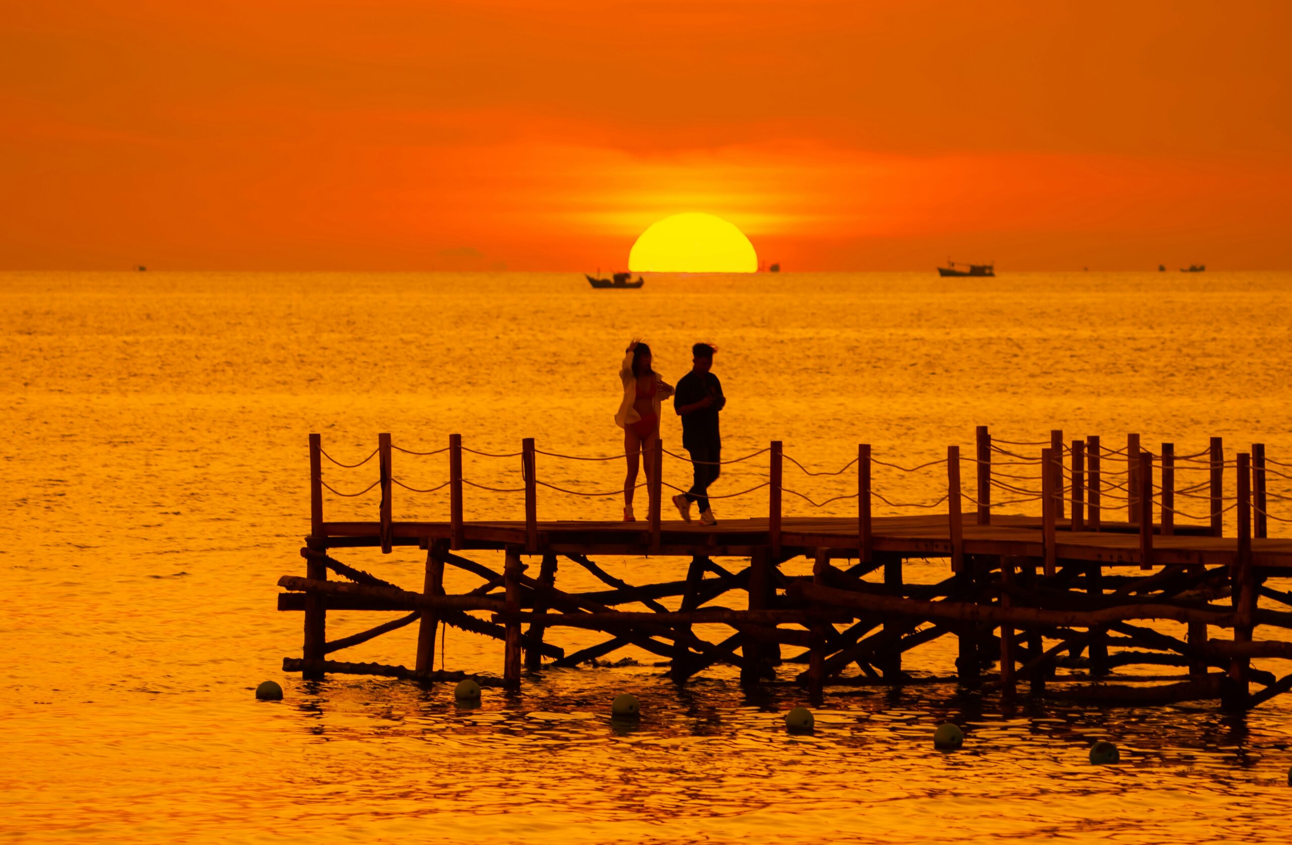 Silhouette of a romantic couple walking on a rustic wooden pier during a breathtaking golden-orange sunset on their Vietnam Honeymoon.